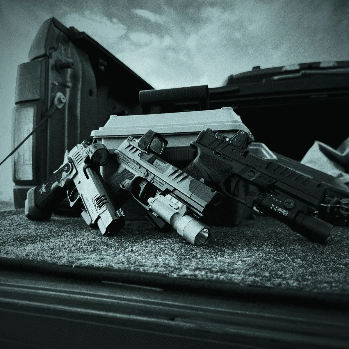 Handguns and a rifle fitted with red dot sights and weapon lights, laid out on a truck bed